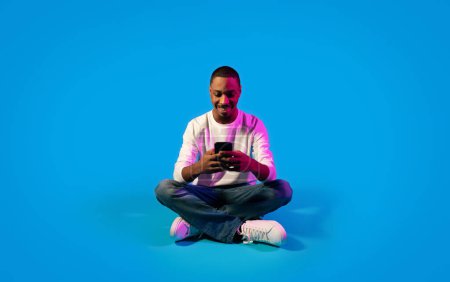 Foto de Cheerful happy young black guy in casual using modern cell phone and smiling over blue background in neon light, chatting with ladies on dating app, surfing on Internet. Gadget addiction concept - Imagen libre de derechos