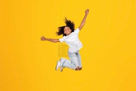 Photo for Cheerful curly teen black schoolgirl in white t-shirt has fun, jumping, freezes in air, enjoy freedom, isolated on yellow background, studio. Lifestyle, study and victory, holidays ad and offer - Royalty Free Image