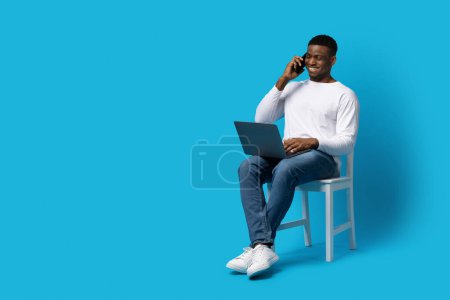 Photo for Modern technologies in business concept. Cheerful handsome young black guy in casual businessman sitting on chair over blue background, have phone conversation and using laptop, copy space - Royalty Free Image