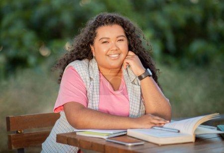 Téléchargez les photos : Formation universitaire. Etudiante hispanique joyeuse apprenant en plein air, posant avec des livres faisant son projet d'étude et ses devoirs assis à table dans le parc à l'extérieur, souriant à la caméra - en image libre de droit