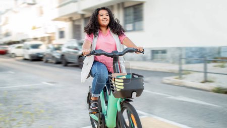Photo for Easy City Transportation. Cheerful Latin Overweight Woman Riding Bicycle On Her Way To College. Lady Enjoying Bike Ride Carrying Books In Basket Posing On A Street In Town Outside. Panorama - Royalty Free Image