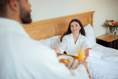 Photo for Morning Surprise. Loving Husband Giving Tray With Breakfast To Cheerful Wife While She Relaxing In Hotel Bedroom Interior. Couple Sharing Joy Of Romantic Honeymoon Vacation. Selective Focus - Royalty Free Image