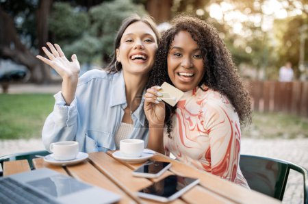 Photo for Easy Payments Concept. Two Happy Women Showing Credit Card While Sitting In Cafe Outdoors, Cheerful Multiethnic Female Friends Paying For Coffee And Laughing At Camera, Closeup Shot - Royalty Free Image