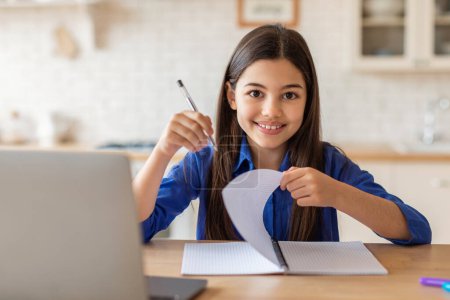 Foto de E-Learning Self Discipline. Happy School Kid Girl At Laptop Taking Notes In Textbook, Sitting At Desk Smiling To Camera At Home (en inglés). Colegiala con éxito de aprendizaje en línea Excelencia en estudios remotos - Imagen libre de derechos