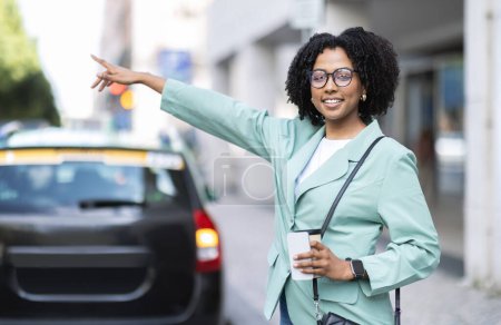 Foto de Feliz hermosa mujer negra joven con traje formal, anteojos coger un taxi en la calle, sosteniendo la taza de papel con café y teléfono inteligente, trabajador que va a la oficina por la mañana, espacio para copiar - Imagen libre de derechos