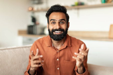 Téléchargez les photos : Portrait d'un homme indien souriant parlant en regardant la caméra et en faisant des gestes, tout en communiquant avec les clients en ligne assis sur le canapé à la maison. Vue de la caméra Web du gars bavarder à distance via un appel vidéo - en image libre de droit