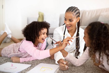 Photo for Family Joy And Fun. Joyful Black Mommy And Daughters Drawing And Fooling Painting Each Others Faces At Home, Lying On Floor In Modern Living Room. Preteen Girls And Mother Painting Together - Royalty Free Image
