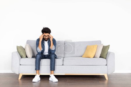 Photo for Unhappy pensive handsome bearded middle eastern young man in smart casual experiencing problems in life, sitting alone on couch in living-room at home, touching his head, looking down, copy space - Royalty Free Image