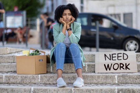Photo for Upset unemployed jobless young black woman sitting on stairs outdoors by need work sign placard, cardbox with office stuff belongings next to her, copy space. Sad lady lost her job - Royalty Free Image
