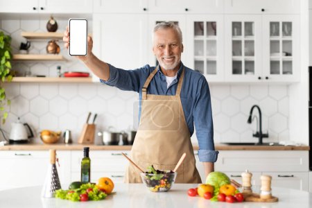 Foto de Aplicación de dieta. Hombre mayor sonriente sosteniendo Smartphone en blanco mientras cocina en la cocina en casa, feliz caballero anciano recomendando aplicación móvil para una nutrición saludable y pérdida de peso, burla - Imagen libre de derechos