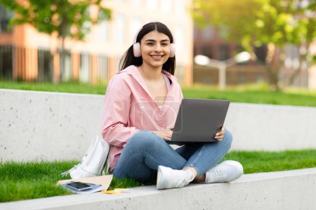 Téléchargez les photos : Heureuse étudiante hispanique dame dans les écouteurs sans fil étudier avec ordinateur portable à l'extérieur, assis avec ordinateur sur le campus, regardant et souriant à la caméra, profiter de l'apprentissage à distance, copier l'espace - en image libre de droit