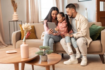 Family Gadgets. Korean parents and baby daughter using digital tablet sitting together on couch in living room indoors. Cheerful father, mother and child websurfing on pad at home