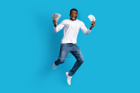 Photo for Cashback, loan, giveaway. Rich wealthy happy handsome young black guy in casual outfit jumping in the air with money cash dollar banknotes in his hands over blue studio background, copy space - Royalty Free Image