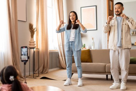 Photo for Family Blogging. Joyful korean parents and kid daughter broadcasting while having fun, shooting video on smartphone at home. Family filming new vlog talking to phone camera, pointing at themselves - Royalty Free Image