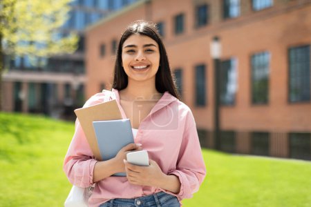 Photo for Portrait of happy student girl with cellphone and workbooks walking in college campus and smiling at camera, having break after classes outdoors, copy space - Royalty Free Image