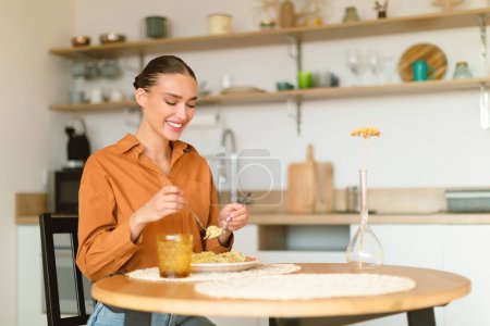 Foto de Mujer caucásica joven comiendo pasta sabrosa, almorzando y disfrutando de deliciosos espaguetis caseros mientras está sentado en la mesa en el acogedor interior de la cocina, espacio de copia gratis - Imagen libre de derechos
