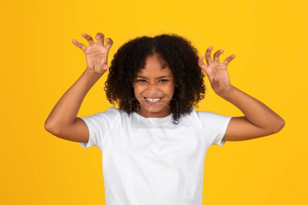 Téléchargez les photos : Joyeux drôle jeune écolière noire bouclée en t-shirt blanc faisant signe de patte de bête, attaque, isolé sur fond jaune, studio. Jouer au jeu, imagination, style de vie, apprentissage, vacances, annonce et offre - en image libre de droit