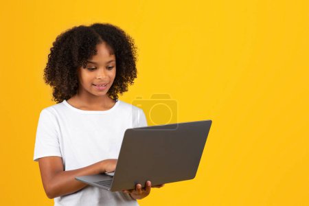 Photo for Cheerful curly teenager black schoolgirl in white t-shirt uses laptop, isolated on yellow background, studio. Study, knowledge remotely, video lesson, social networks, ad and offer - Royalty Free Image