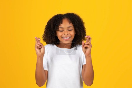 Photo for Cheerful curly teenager black schoolgirl in white t-shirt crossed fingers, make wish, isolated on yellow background, studio. Hope, faith, exam expects, good news, ad and offer - Royalty Free Image