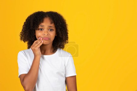 Photo for Sad curly teenager black schoolgirl in white t-shirt presses hand to cheek, suffering from toothache, isolated on yellow background, studio. Caries, health problems, dentistry, ad and offer - Royalty Free Image