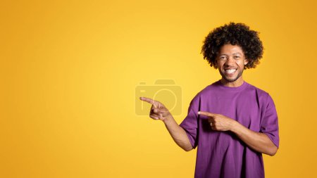 Téléchargez les photos : Glad black adulte bouclé en t-shirt violet pointe les doigts vers l'espace vide, isolé sur fond orange, studio, panorama. Recommandation, conseils, vente et bonnes nouvelles, annonce et offre - en image libre de droit