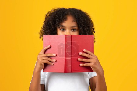 Téléchargez les photos : Sérieux smart curly adolescente noire écolière en t-shirt blanc regarde par derrière livre, isolé sur fond jaune, studio, gros plan. Éducation, études, loisirs et devoirs - en image libre de droit
