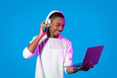 Téléchargez les photos : Joyeux jeune homme noir barbu heureux en utilisant un ordinateur portable moderne et des écouteurs sans fil, en appuyant sur le bouton sur le casque et en souriant sur fond bleu dans la lumière au néon, avoir un chat vidéo, copier l'espace - en image libre de droit