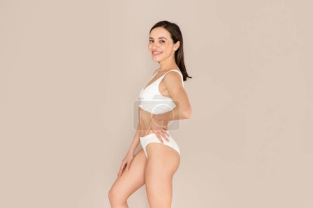 Photo for Healthy body. Cheerful stunning pretty young lady posing on beige studio background, wearing white comfortable underwear, smiling at camera, showing perfect body, side view, copy space - Royalty Free Image