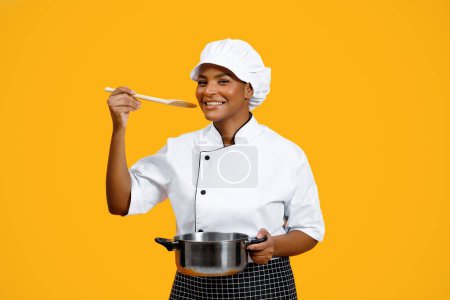 Photo for Black chef lady with saucepan and spoon tasting food over yellow background, smiling african american female cooker in toque preparing meal and looking at camera, enjoying cooking, copy space - Royalty Free Image