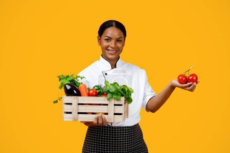 Téléchargez les photos : Femme noire souriante tenant une caisse en bois avec des légumes biologiques et regardant la caméra, heureuse cuisinière afro-américaine démontrant des ingrédients, isolée sur fond jaune - en image libre de droit