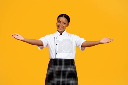 Téléchargez les photos : Chef noir souriant femme chef ouvrant les bras à la caméra, heureuse cuisinière afro-américaine dame présentant quelque chose sur ses paumes ouvertes, saluant quelqu'un, geste de bienvenue tout en posant sur fond jaune - en image libre de droit