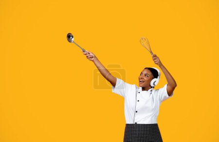 Foto de Mujer Chef Negro feliz en los auriculares bailando en el fondo del estudio amarillo, alegre cocinero afroamericano señora sosteniendo los utensilios y señalando hacia fuera en el espacio de copia, haciendo que Dab se mueva, divirtiéndose - Imagen libre de derechos