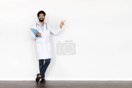 Téléchargez les photos : Beau sourire joyeux jeune barbu médecin du Moyen-Orient en vêtements de travail avec stéthoscope et tableau médical montrant l'espace de copie sur fond de mur blanc, bannière web. Soins de santé, concept médical - en image libre de droit