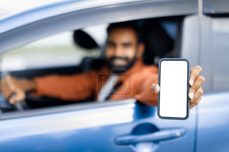 Photo for Navigator Application. Happy Arabic Guy Sitting Inside Car Displaying Smartphone With Empty Screen Through Opened Auto Window. Driver Man Advertising Navigation Mobile App. Selective Focus On Phone - Royalty Free Image