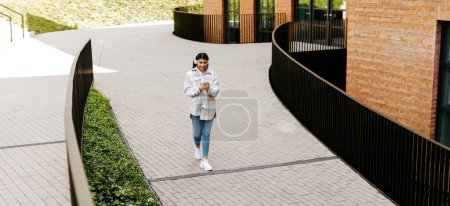 Photo for Teen student lady walking near university building outdoors, wearing headphones and using cellphone, spending free time after classes outside, panorama, free space - Royalty Free Image