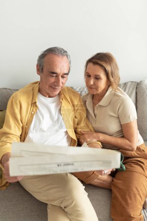 Photo for Loving senior spouses reading newspaper together, sitting on sofa and spending morning at home, aged european couple enjoying moments on weekend - Royalty Free Image