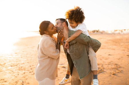 Photo for Loving parents spending time with son outdoors walking on beach, spouses kissing while man piggybacking kid boy, family spending evening by seaside - Royalty Free Image
