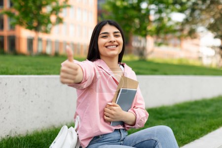 Photo for Excited hispanic student lady gesturing thumbs up and smiling, posing holding books, sitting in college campus or park. I like my college, girl approving modern education concept - Royalty Free Image