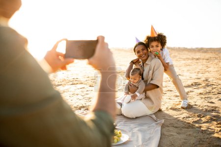 Photo for Happy family resting by seaside at sunset, man taking photo of his wife and children on cellphone, male capturing moments of summer day evening - Royalty Free Image