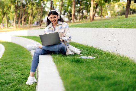 Technology and studentship concept. Cheerful lady student using laptop computer and wearing headphones, typing on keyboard, sitting in park or college campus outdoors