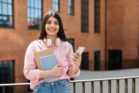 Foto de Retrato de una alegre estudiante latina con teléfono inteligente y libros de trabajo posando al aire libre y sonriendo a la cámara, femenina usando el teléfono mientras está parada afuera cerca de la universidad - Imagen libre de derechos