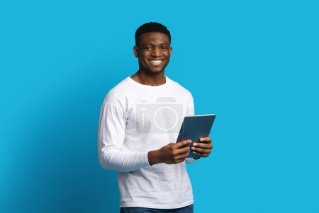 Photo for Cheerful handsome young black guy in casual using modern digital tablet and smiling over blue background, chatting with ladies on dating app, surfing on Internet, copy space. Gadget addiction concept - Royalty Free Image