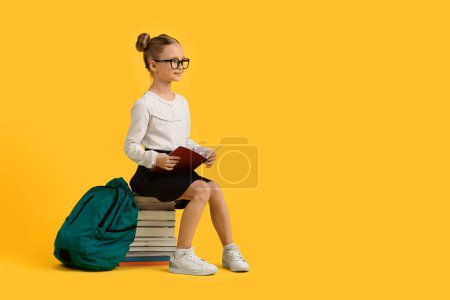 Photo for Study Concept. Smiling Little Schoolgirl Sitting On Pile Of Books And Looking Aside At Copy Space Over Yellow Studio Background, Cute Preteen Female Kid Wearing Eyeglasses And Holding Workbook - Royalty Free Image