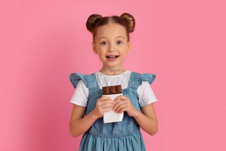 Photo for Portrait Of Cute Little Girl With Chocolate Bar In Hands Looking At Camera, Happy Preteen Female Kid Enjoying Sweet Dessert, Cheerful Child Posing Over Pink Background In Studio, Copy Space - Royalty Free Image