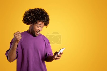 Foto de Glad sorprendido negro adulto rizado chico en púrpura camiseta mira el teléfono inteligente con gesto de victoria y celebra el éxito, aislado sobre fondo naranja, estudio. Grandes noticias, ganar, anuncio y oferta - Imagen libre de derechos