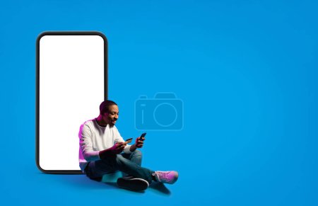Téléchargez les photos : Joyeux beau jeune homme noir assis sur le sol par un téléphone énorme avec maquette sur fond bleu dans la lumière au néon, en utilisant smartphone et tenant la carte de crédit, concept bancaire en ligne, e-commerce, espace de copie - en image libre de droit