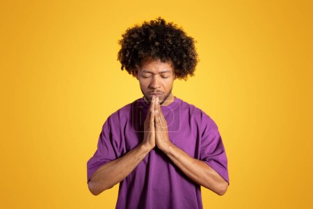 Photo for Calm serious black adult curly man with closed eyes in purple t-shirt prays with hands, isolated on orange background, studio. Dream, make a wish, hope and faith, advertisement and offer - Royalty Free Image
