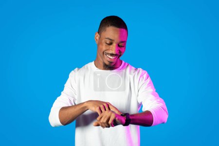 Photo for Happy handsome young black man in neon light looking at modern smartwatch on his hand wrist, blue background, enjoying his new gadget, copy space. Modern technologies in life concept - Royalty Free Image