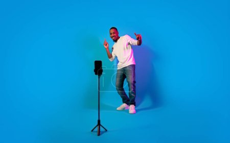 Téléchargez les photos : Concept de blogging. Charismatique joyeux élégant beau jeune homme afro-américain performant devant un téléphone portable sur trépied sur fond bleu, pleine longueur, espace de copie - en image libre de droit