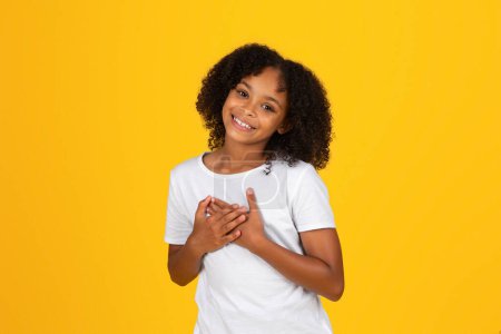 Photo for Satisfied glad curly teenager black girl in white t-shirt presses hands to chest, says thank, isolated on yellow background, studio. Love, gratitude gesture, study, knowledge, ad and offer - Royalty Free Image